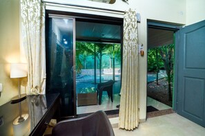 A hotel room with a sliding glass door leading to a pool area, a chair, and a desk with a lamp. - Silvanus Forest Retreat (Alibag)