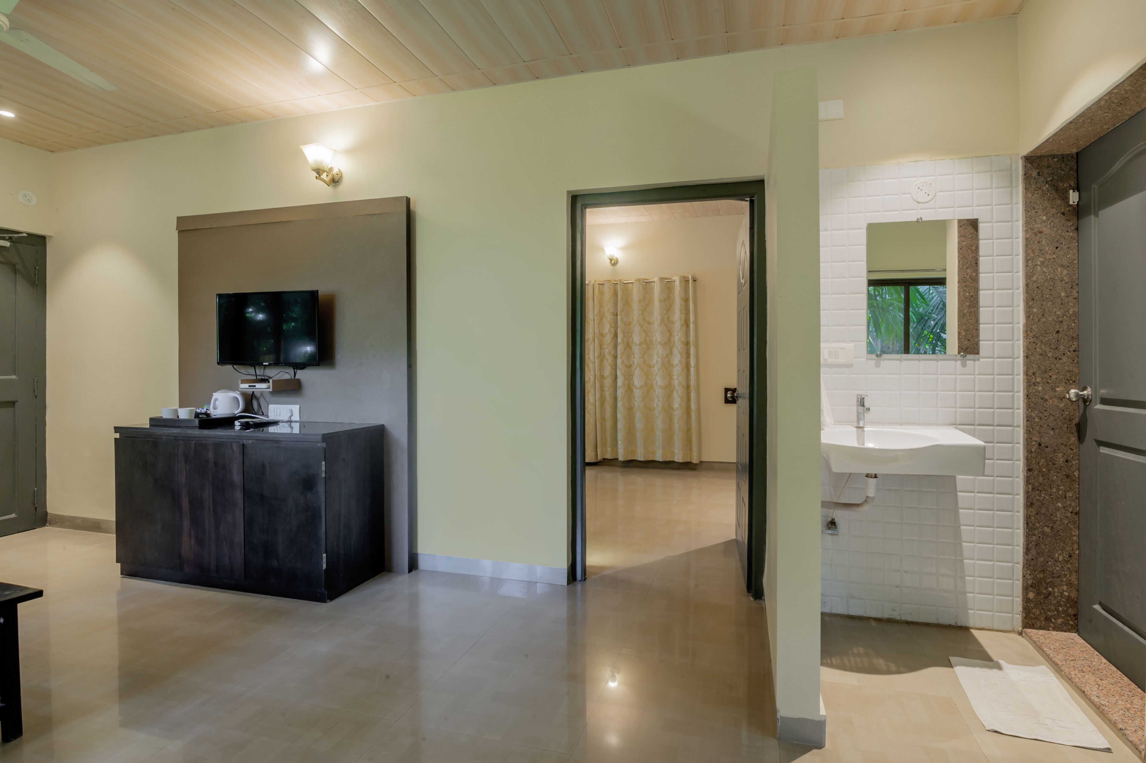 A modern hotel room with a dark wooden desk, a flat-screen TV, a white tiled bathroom with a sink and mirror, and a wooden ceiling.