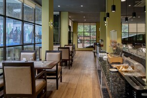 Un restaurante con mesas y sillas de madera, una barra de buffet con distintos alimentos y amplios ventanales que ofrecen vistas al exterior.