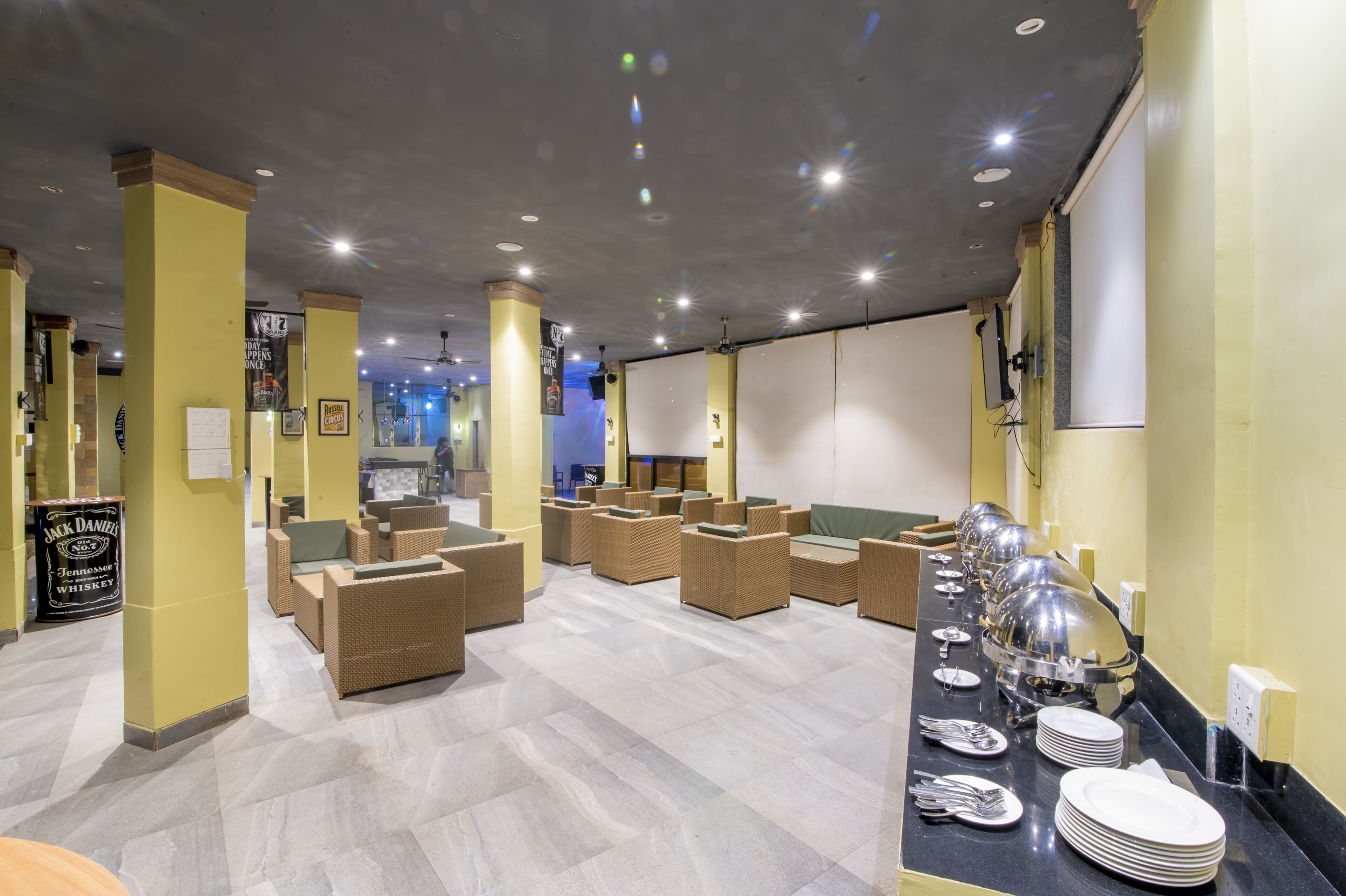 Un hall de hotel con muebles de mimbre, una zona de bufé con platos y cubiertos, y un cartel de Jack Daniel's.