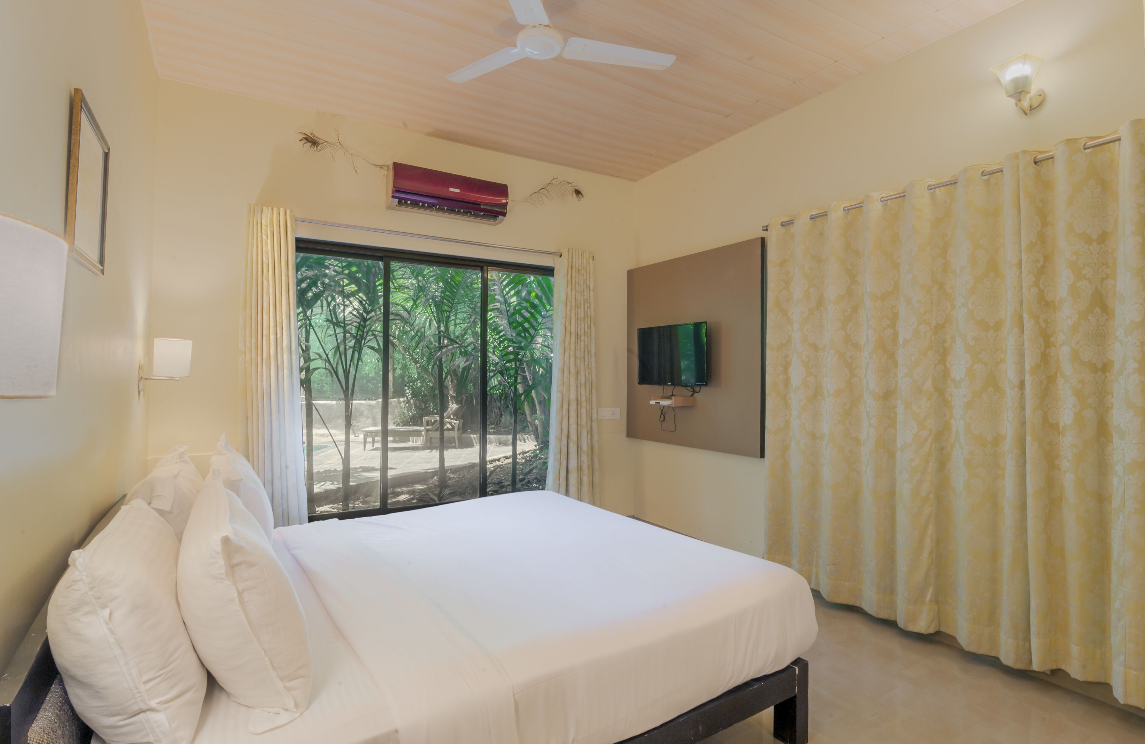 A bedroom with a bed, a ceiling fan, an air conditioning unit, a TV mounted on the wall, and a window with curtains.