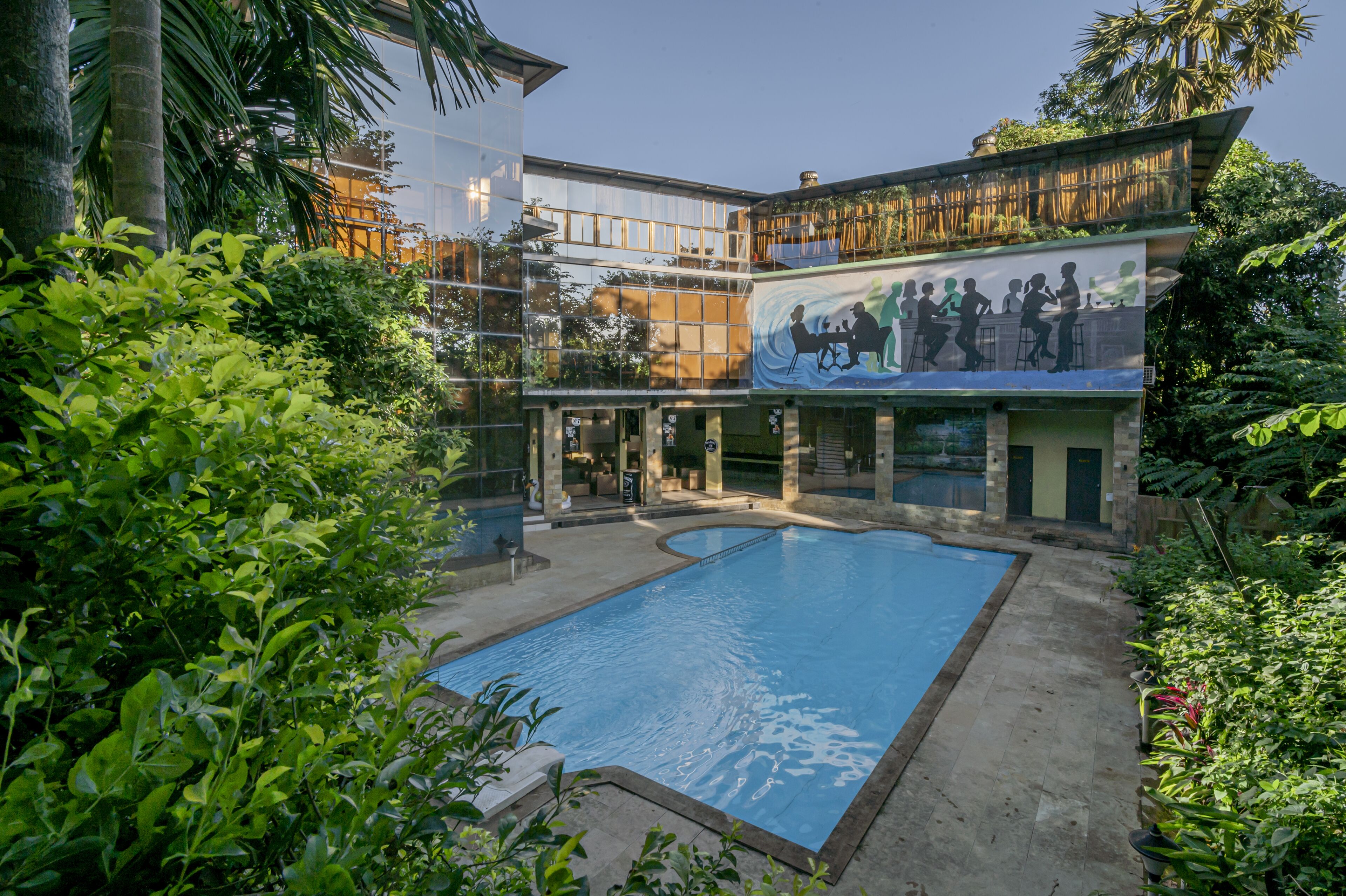 Zona de piscina con un mural, rodeada de abundante vegetación y un edificio de varios pisos con grandes ventanales.