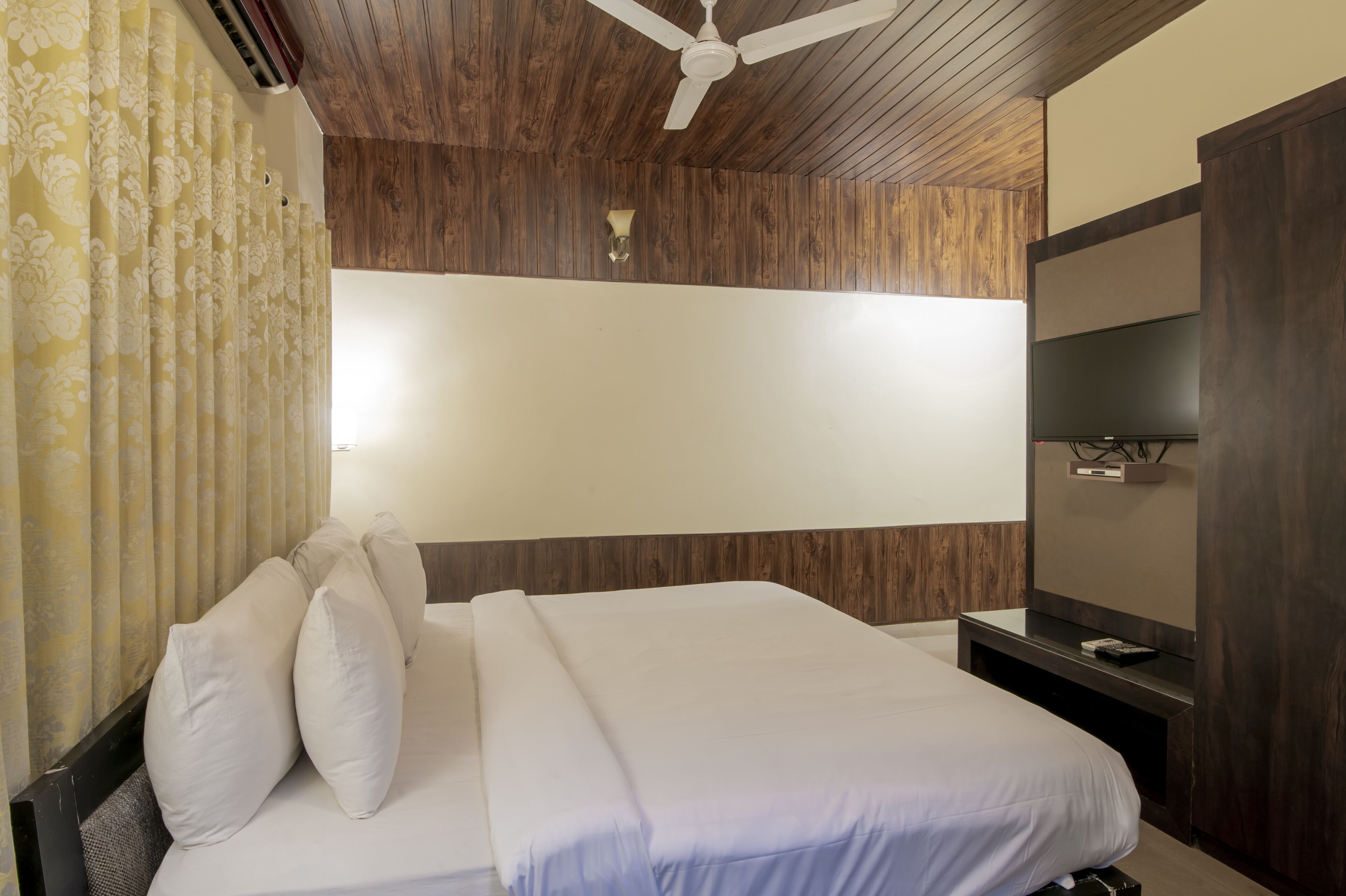 Camera d'albergo con soffitto in legno, letto con biancheria bianca, TV a muro e ventilatore a soffitto.