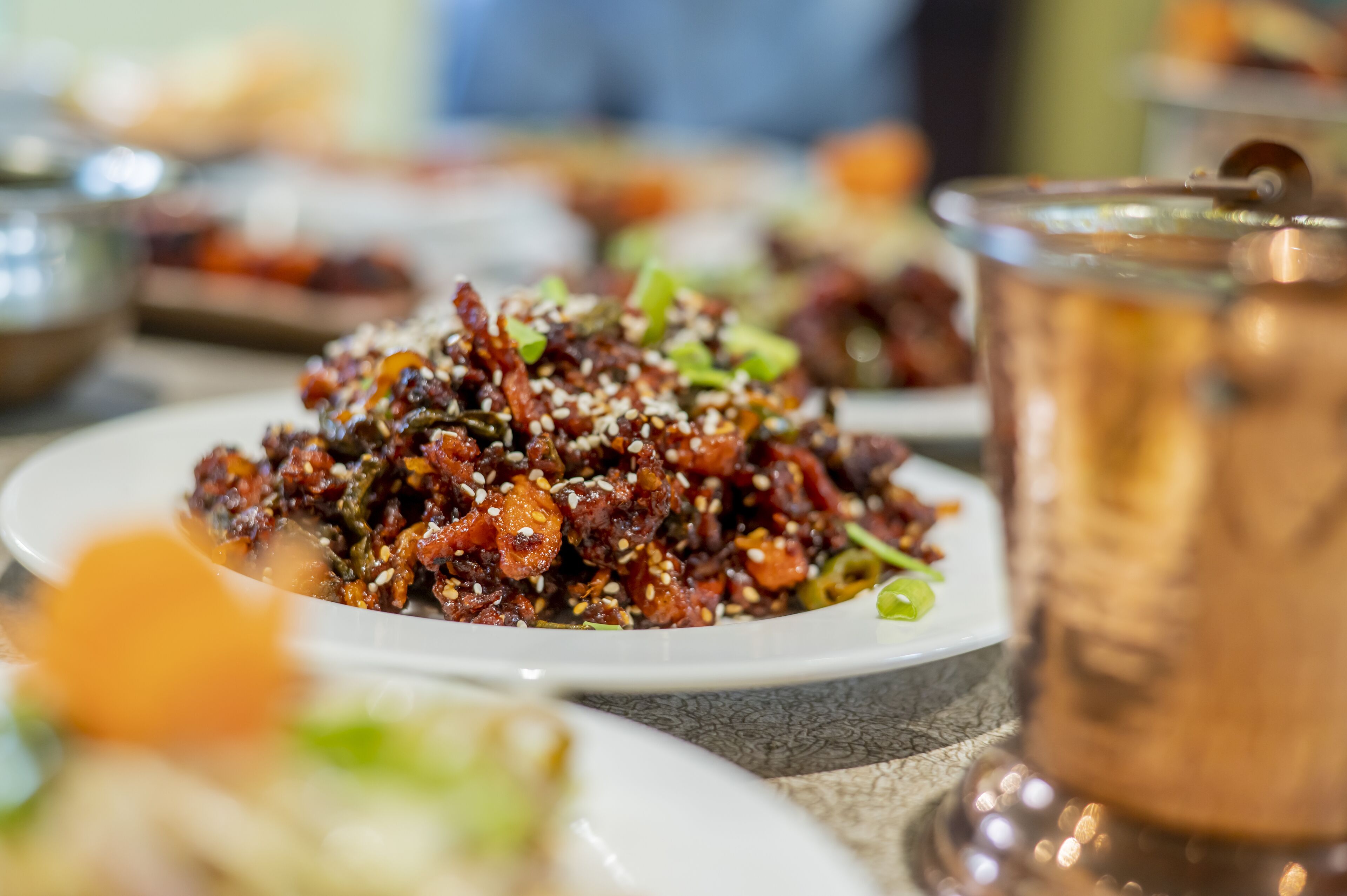A plate of food with sesame seeds and green onions, a glass of beverage, and a blurred background of a dining area.