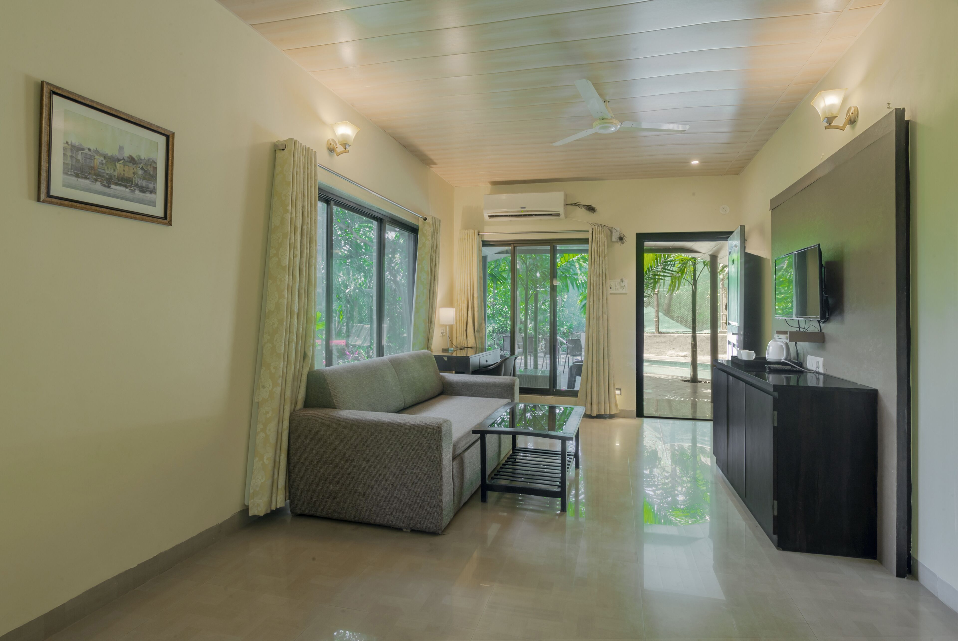 A modern living room with a sofa, a glass coffee table, and a TV on a wooden cabinet. There is a large window with curtains, an air conditioning unit, and a door leading outside.