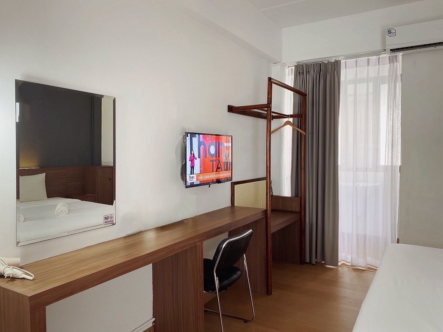 A hotel room with a wooden desk, a chair, a TV mounted on the wall, a bed, and a window with curtains.