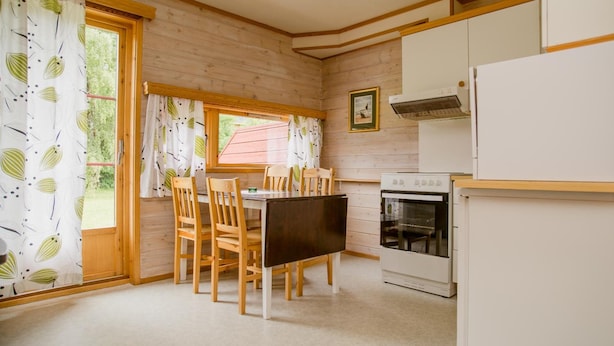 Een kleine keuken met houten kastjes, een eettafel met stoelen, een inbouwoven en een raam met gordijnen.
