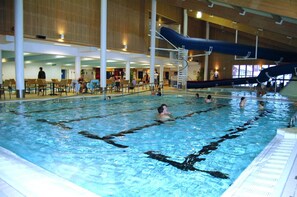 Une piscine intérieure dotée de plusieurs couloirs de nage, d’un toboggan et d’un espace détente avec des tables et des chaises.