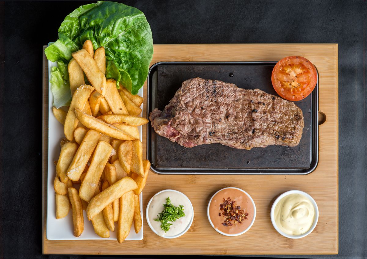 Pommes frites, en grillet bøf, en skive tomat og to dypsaucer på en træplade.