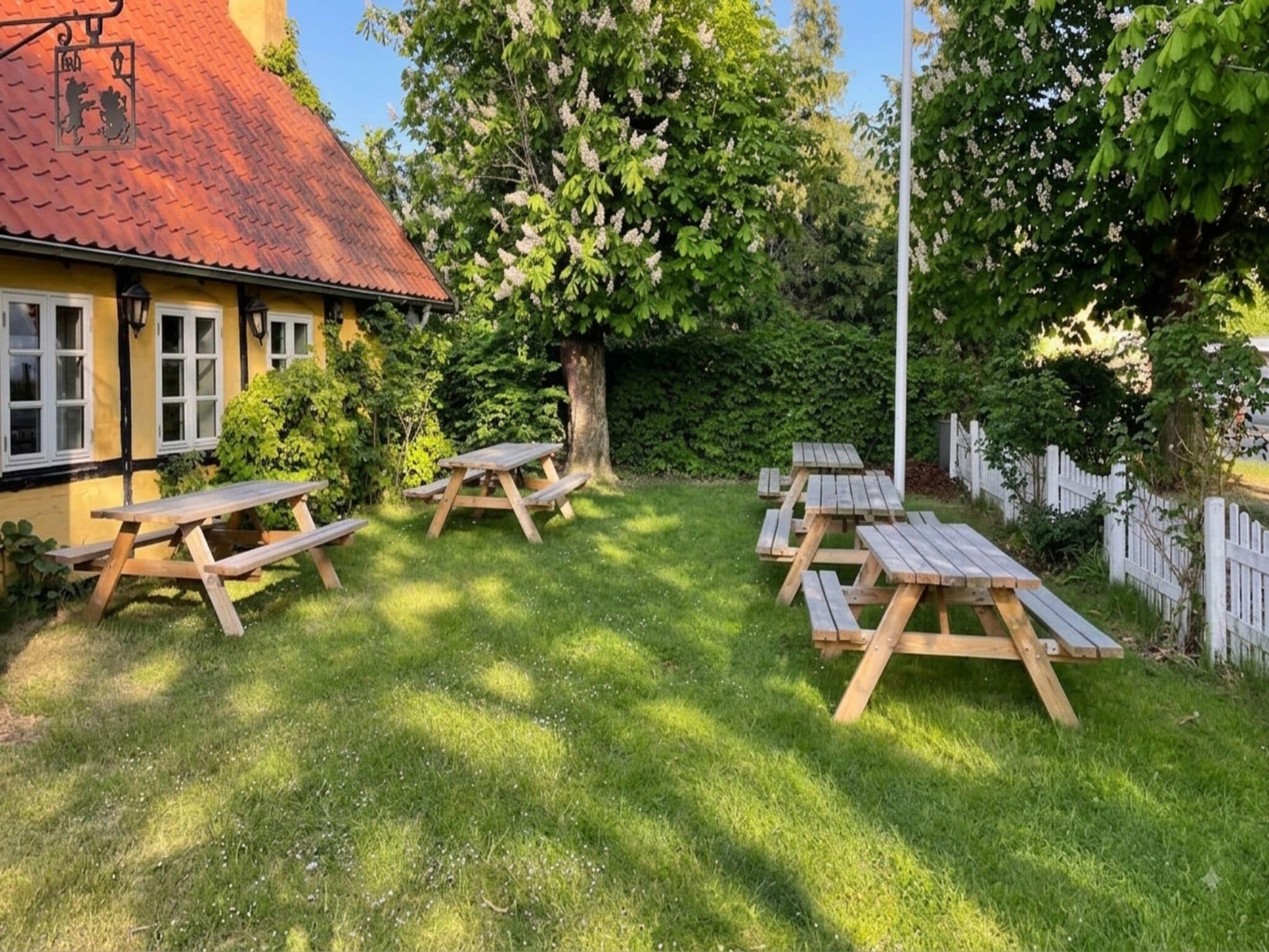 A garden with picnic tables arranged in a semi-circle, surrounded by lush greenery and a building with a red roof.