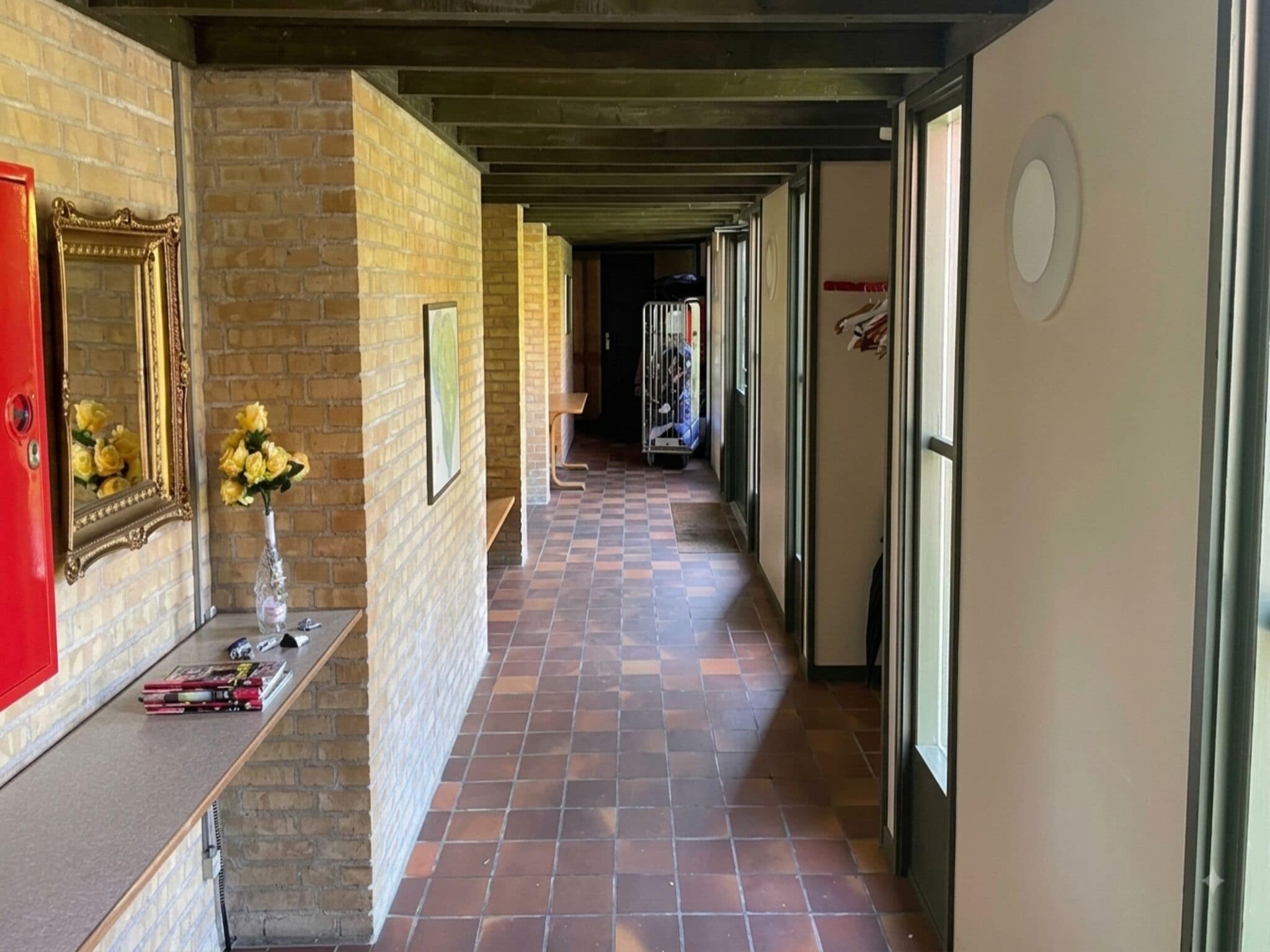 A corridor with a tiled floor, a brick wall, a mirror with a vase of flowers, and a door with a coat rack.