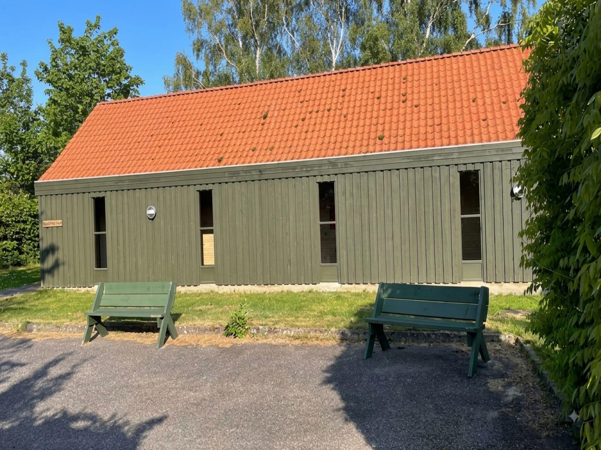 A building with a red-tiled roof and green siding, two green benches in front, and trees in the background.