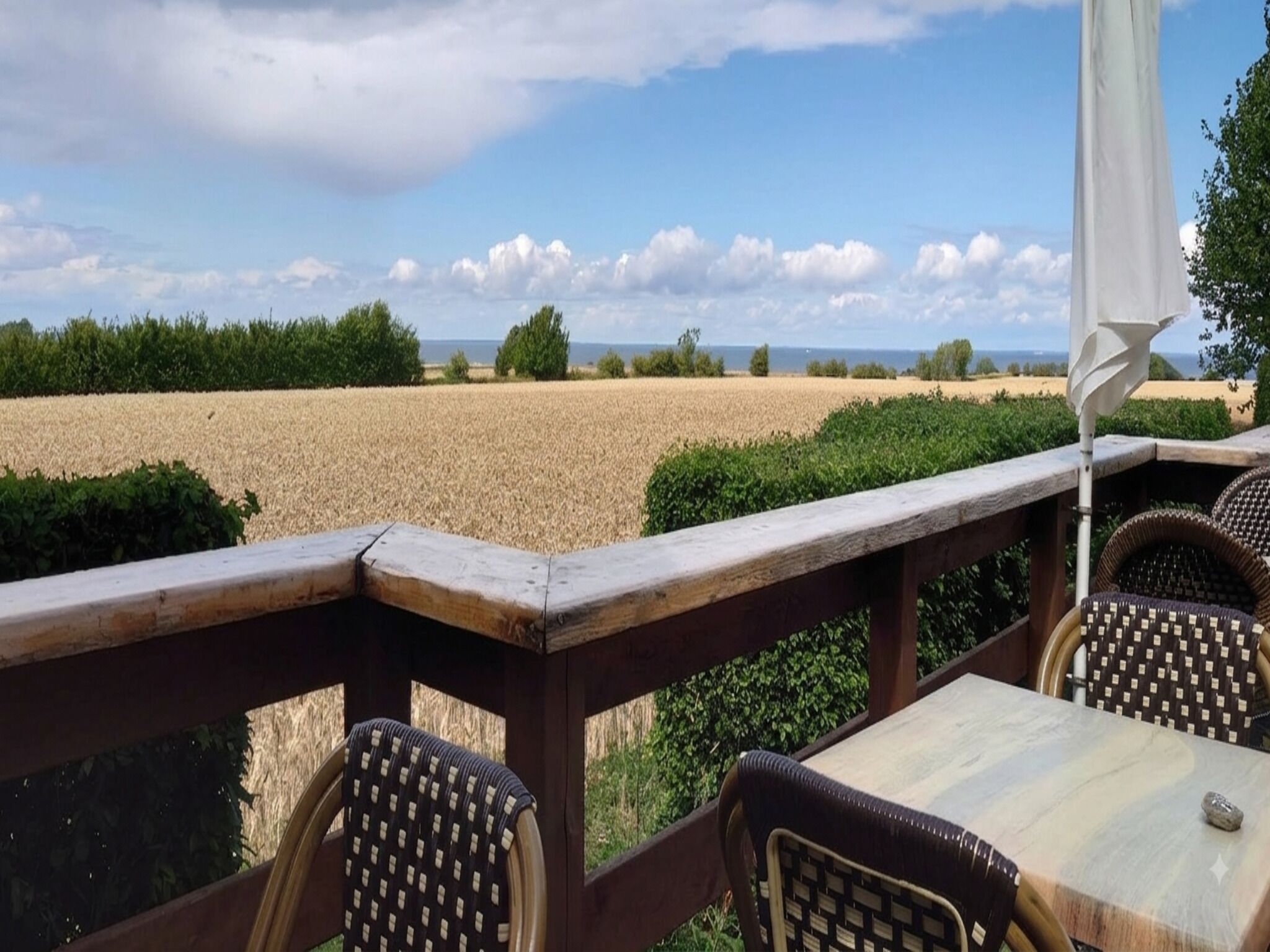 En terrasse med udsigt over et kornfelt, et gelænder af træ og fletstole.
