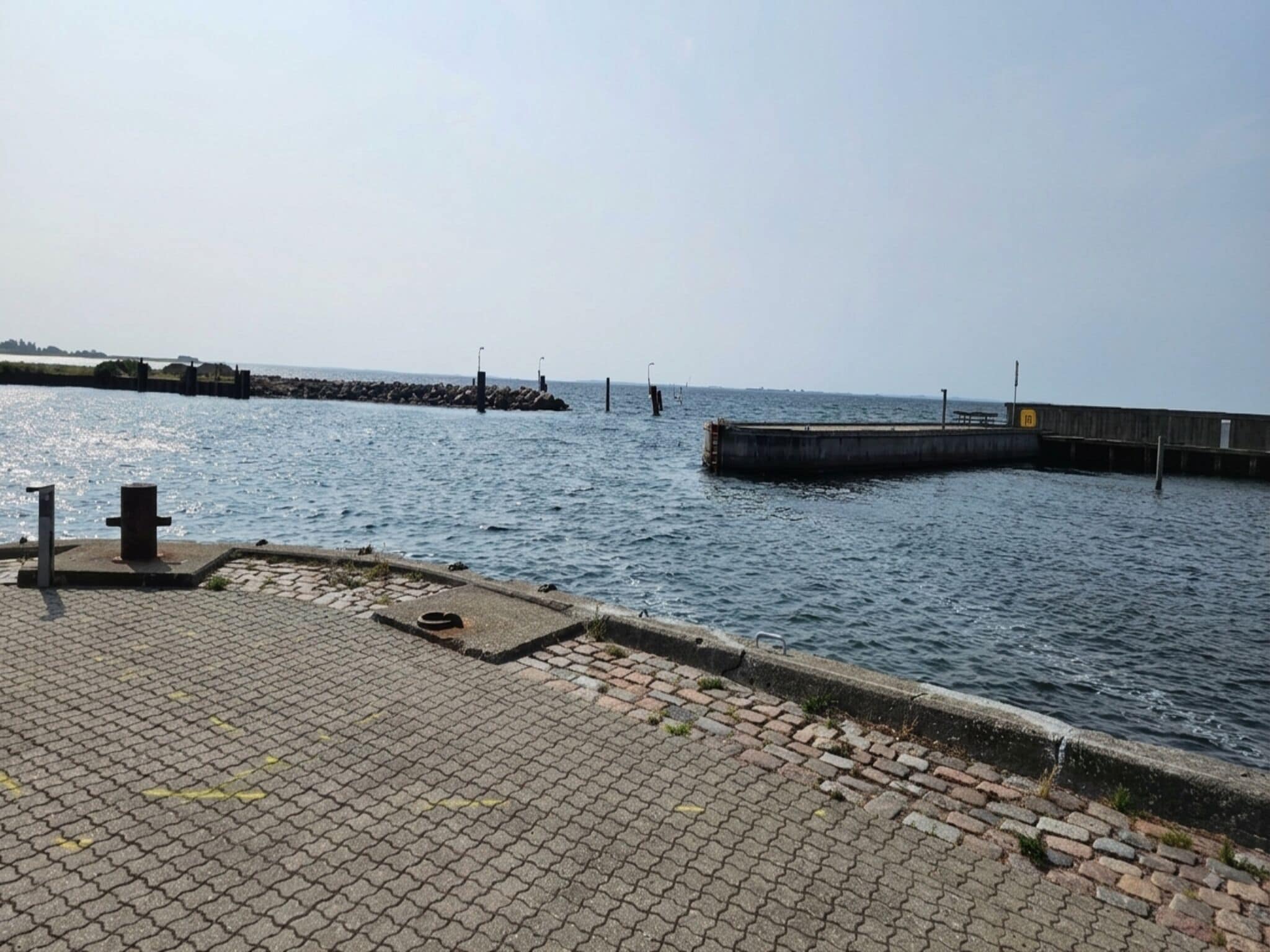 A waterfront promenade with a paved walkway, a wooden pier extending into the water, and a distant shoreline.