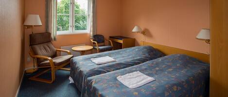 A hotel room with two beds, a desk, two chairs, a window, and two lamps.
