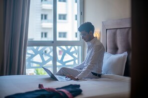 A person sitting on a bed with a laptop, a window with a view of buildings, and a headboard. - Hôtel Étoile du Nord (Tangier)