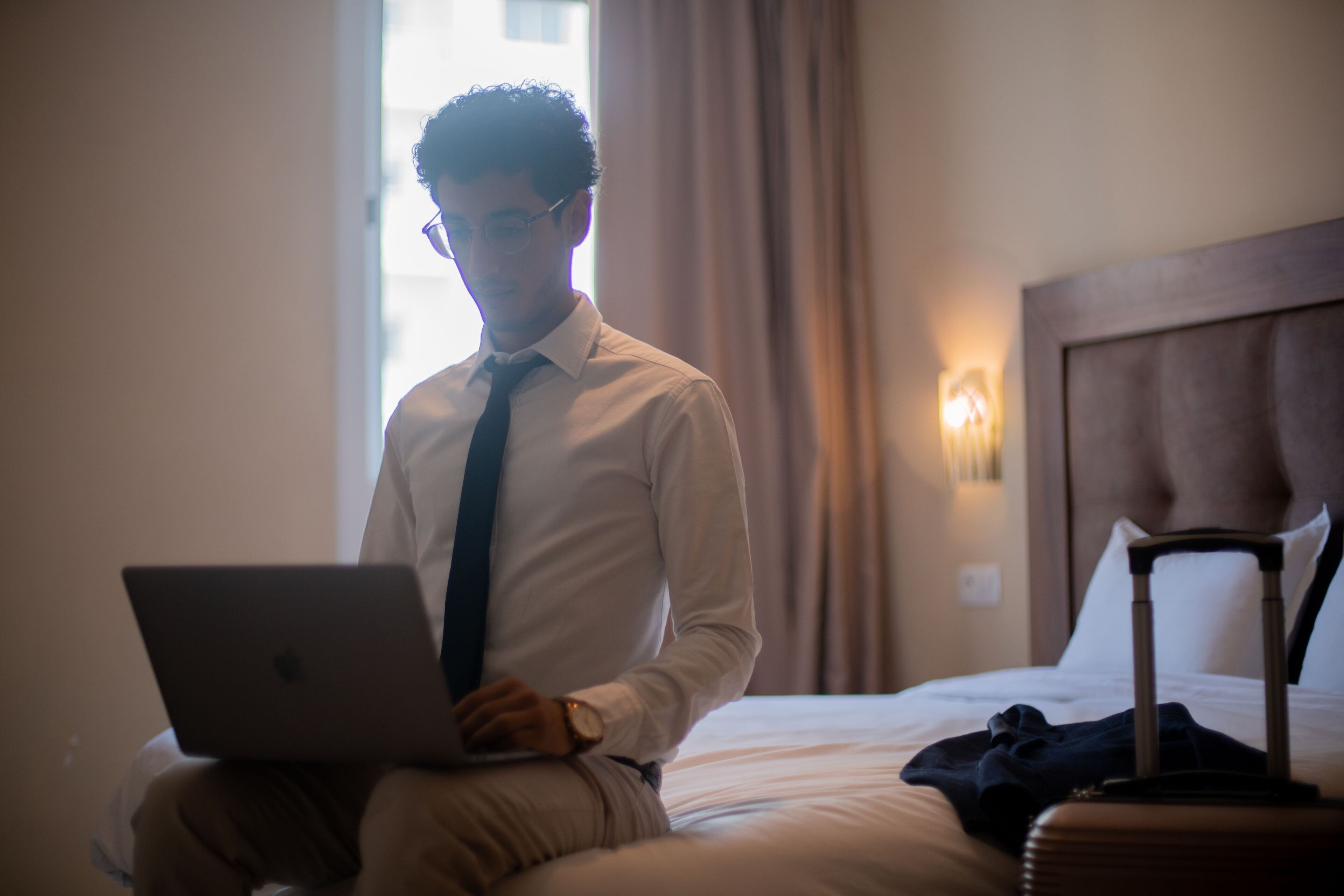 Een persoon zittend op een bed met een laptop, een koffer en een kussen.