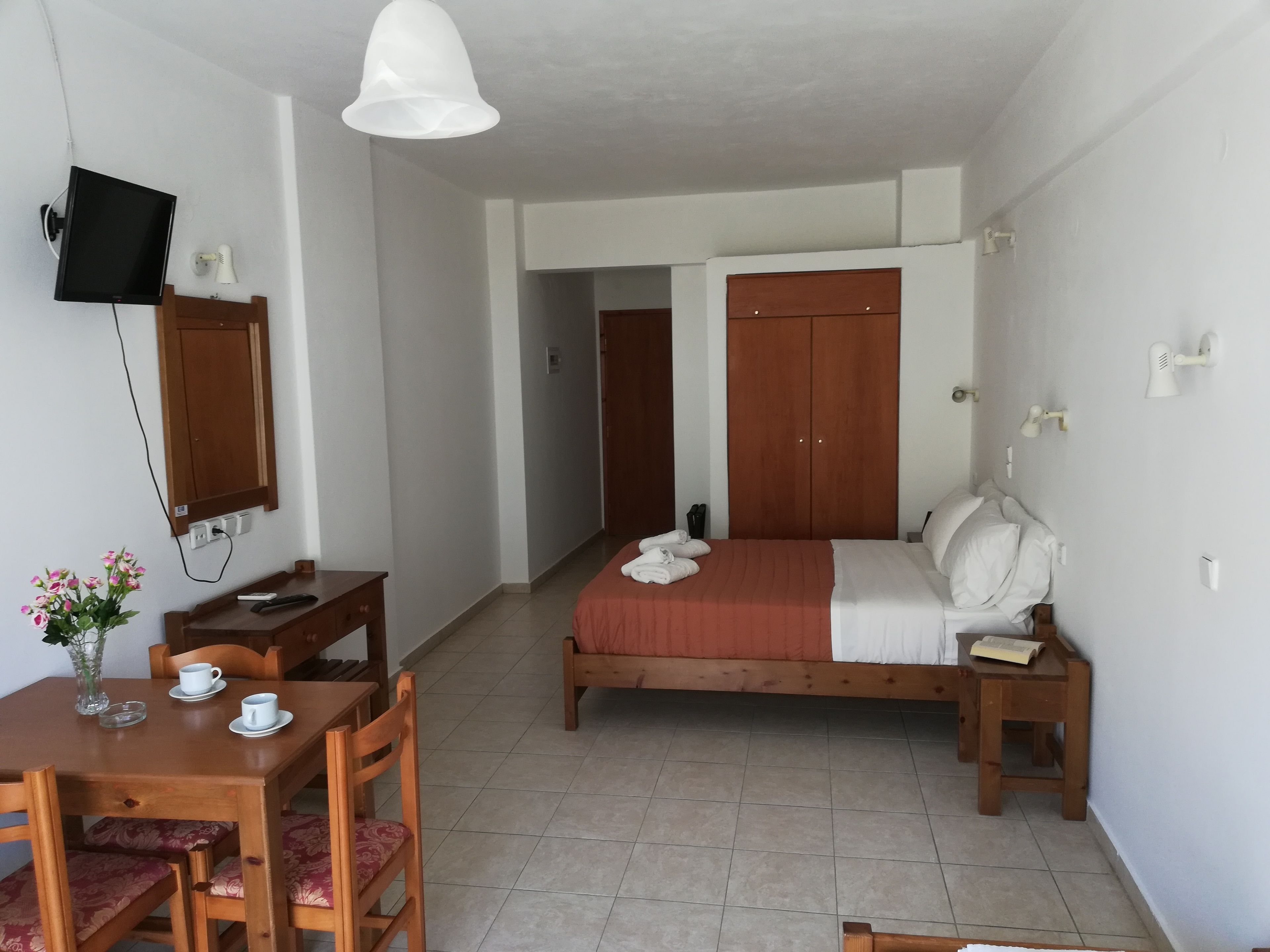 A hotel room with a bed, bedside table, a desk with chairs, a TV mounted on the wall, and a ceiling light.