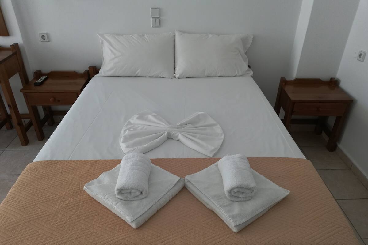 Un lit bien fait, avec des draps blancs, un grand oreiller en forme de nœud et deux serviettes enroulées posées sur une couverture matelassée marron.