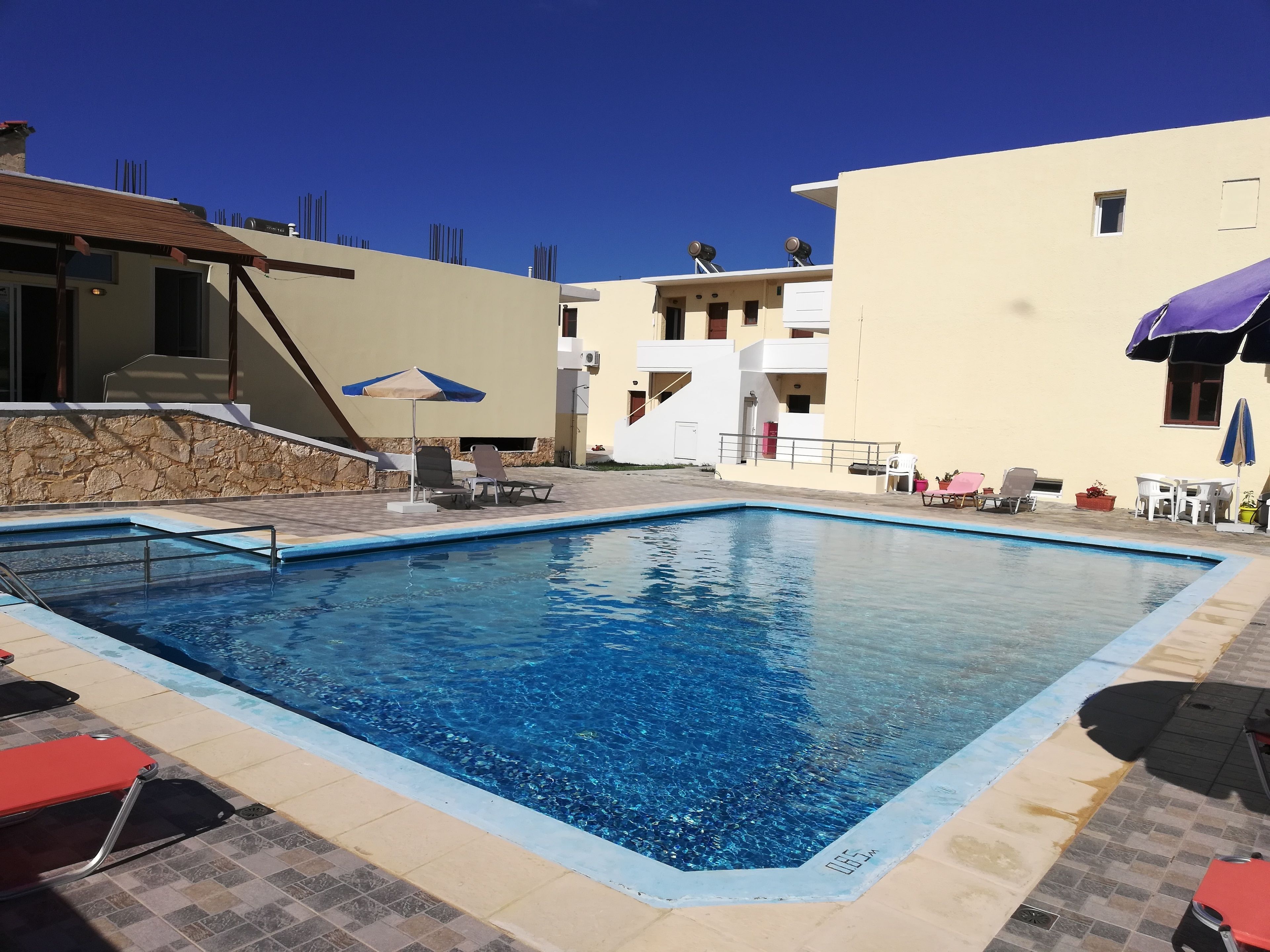 A swimming pool with lounge chairs and umbrellas, surrounded by buildings with balconies and a stone wall.