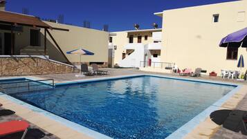 A swimming pool with lounge chairs and umbrellas, surrounded by buildings with balconies and a stone wall.