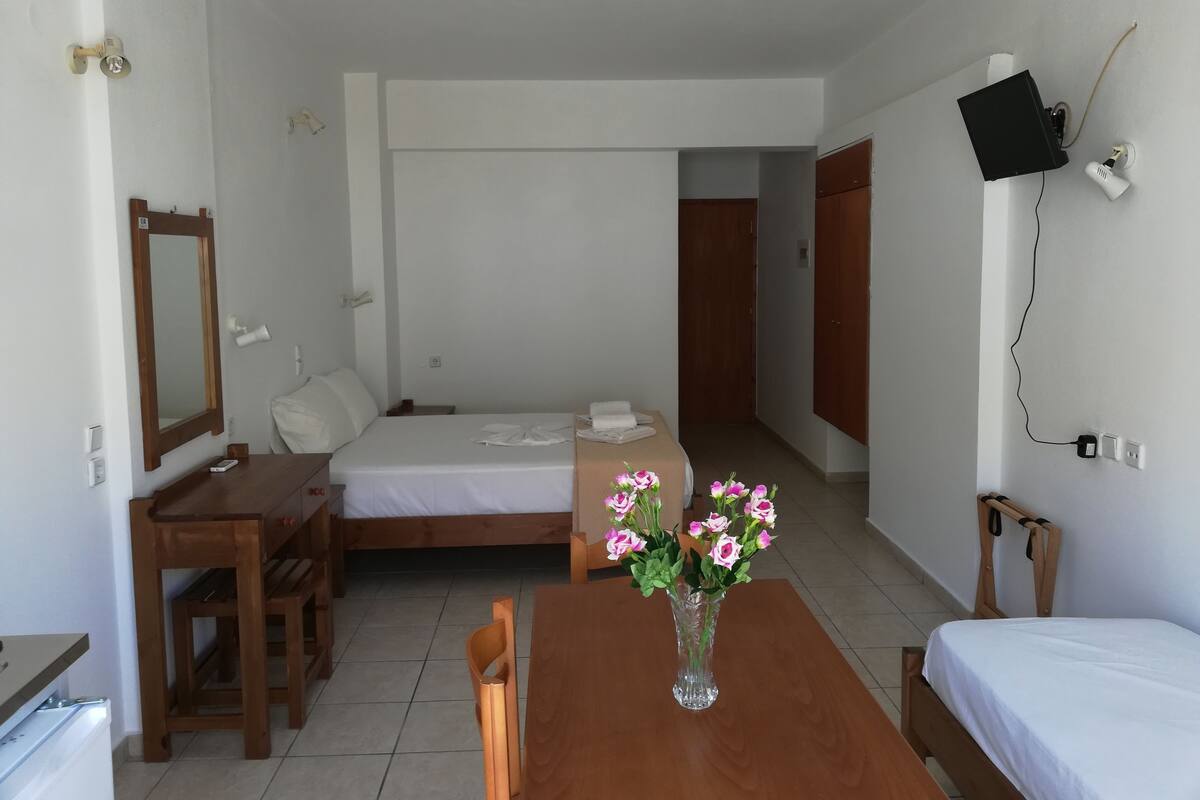 Une chambre d’hôtel comprenant un lit, un bureau, une chaise, une table avec un vase de fleurs, un téléviseur fixé au mur et une porte donnant sur une autre pièce.
