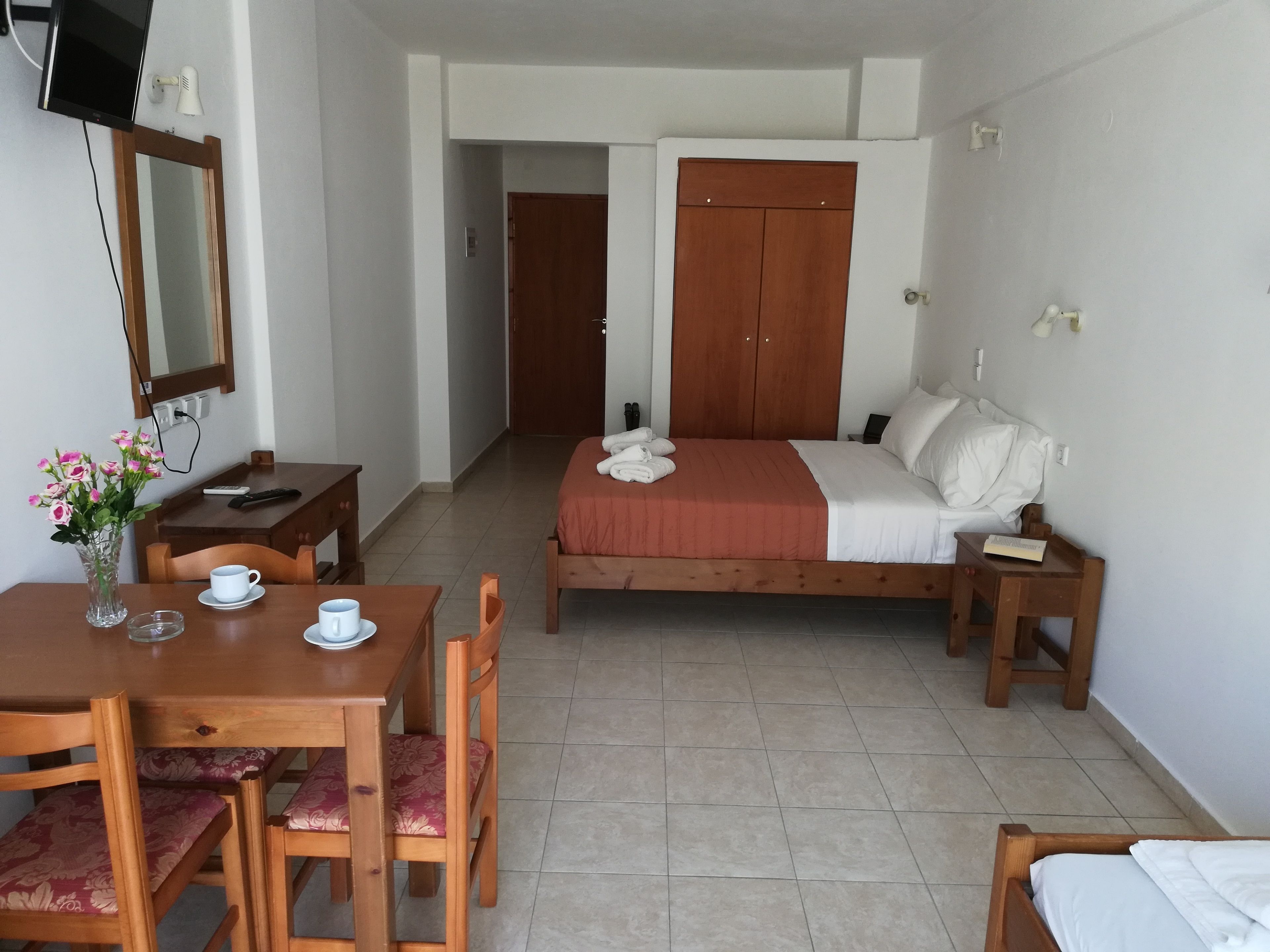 Una camera d'albergo con un letto in legno, un comodino in legno, un tavolino da notte, una scrivania in legno con una sedia, un piccolo tavolo con una tazza e un piattino, e un vaso con fiori.