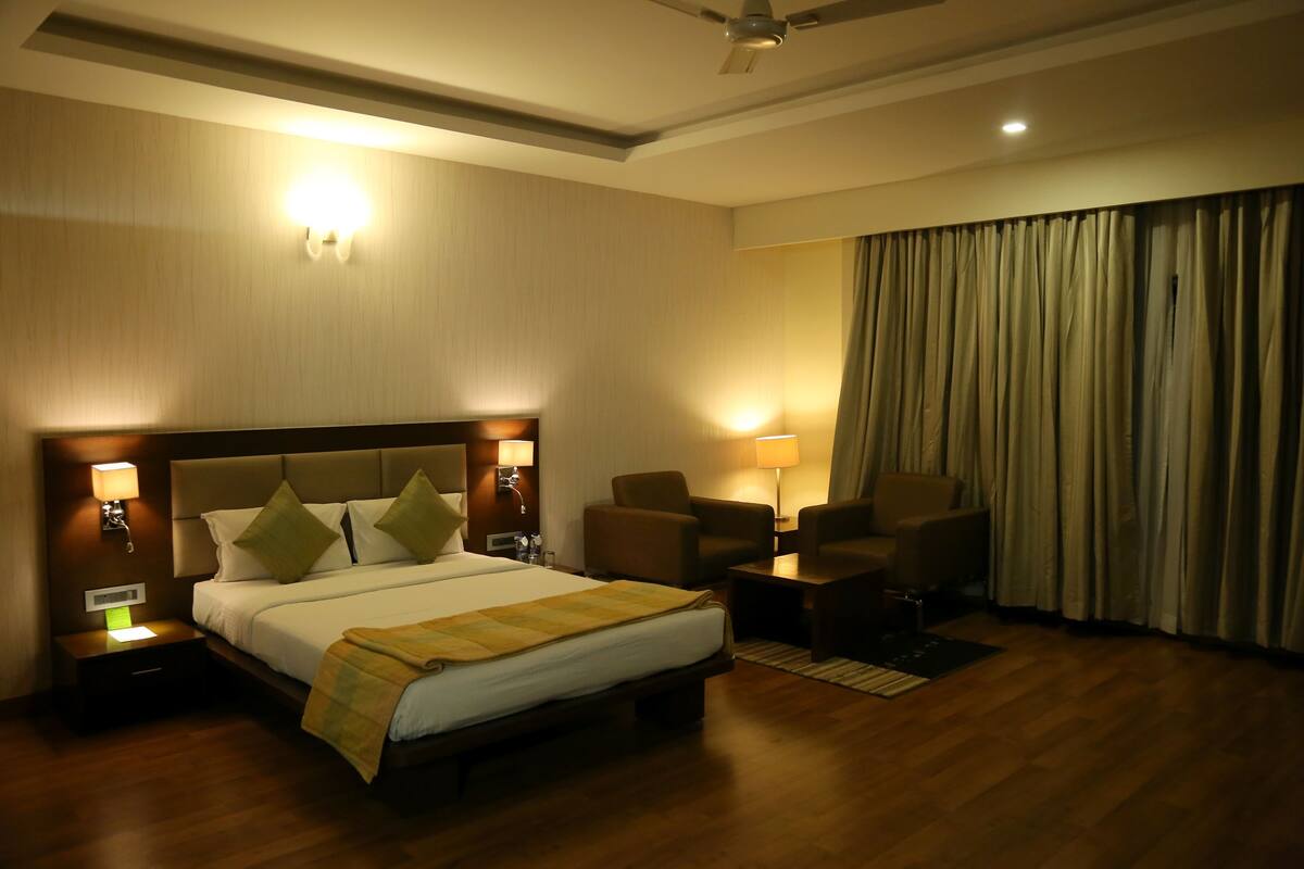 A hotel room with a bed, two chairs, a desk, a ceiling fan, and a large window with curtains.