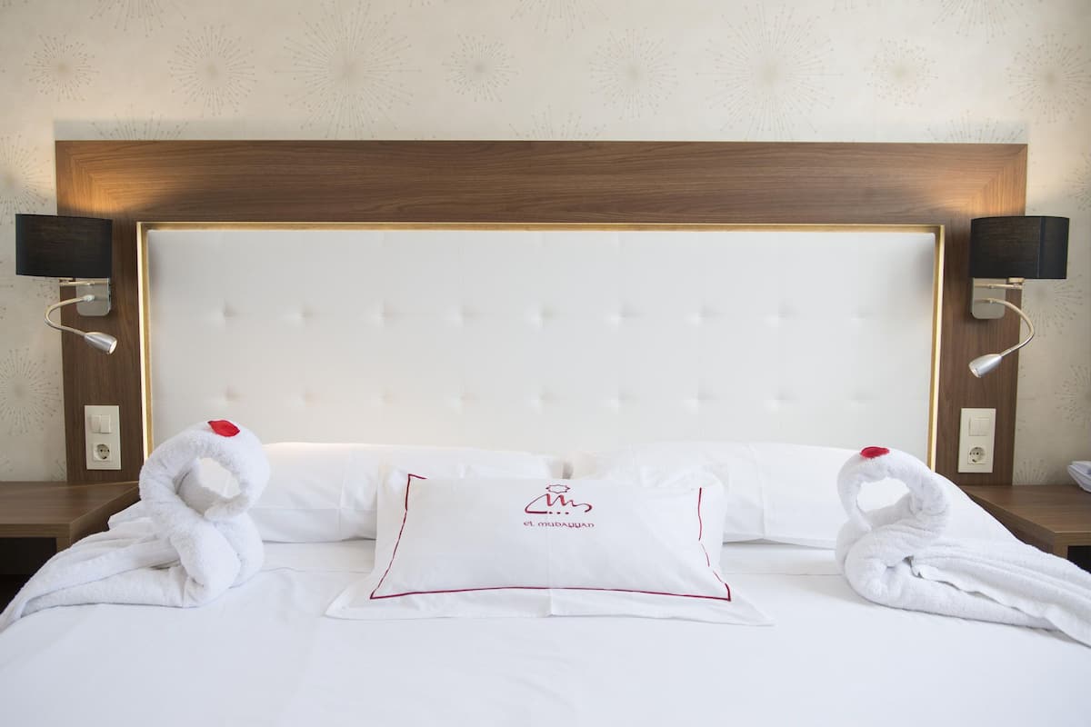Habitación de hotel con cabecera capitonada, ropa de cama blanca y dos toallas con forma de cisnes con detalles rojos.