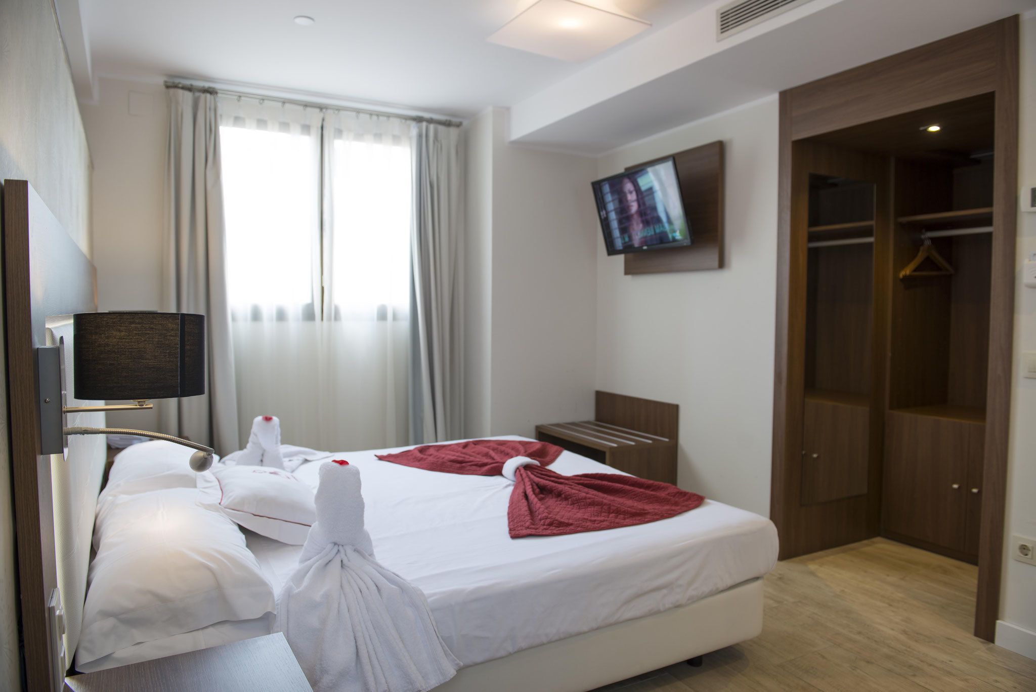 A hotel room with a bed, a television mounted on the wall, a wardrobe, and a window with curtains.