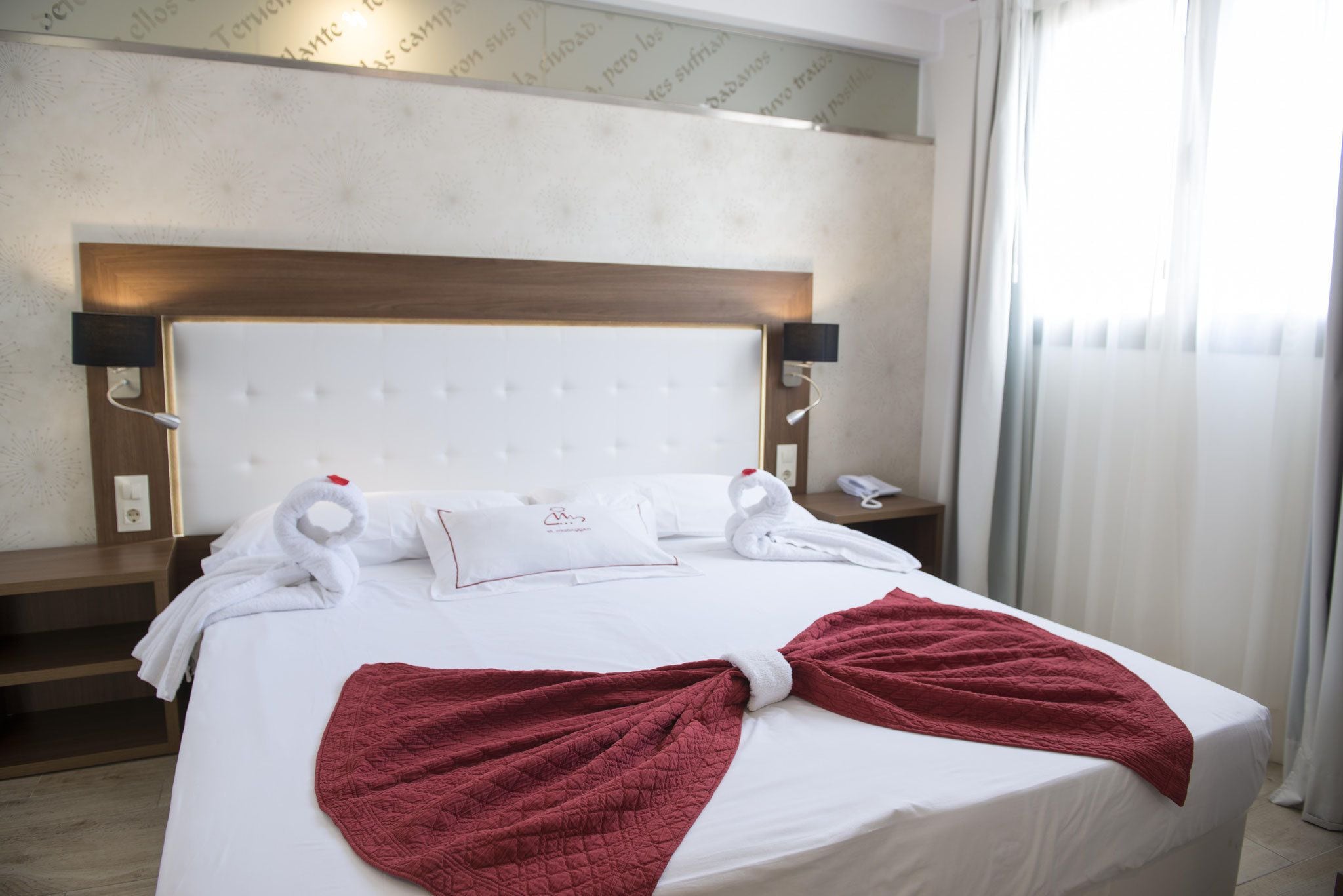 A hotel room with a large bed, a wooden headboard, two bedside tables with lamps, and a window with curtains.