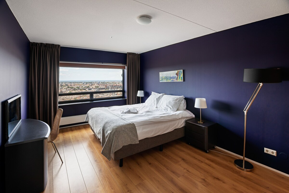Una habitación de hotel moderna con una cama grande, un escritorio, una silla, una mesita de noche, una lámpara y un cuadro en la pared.