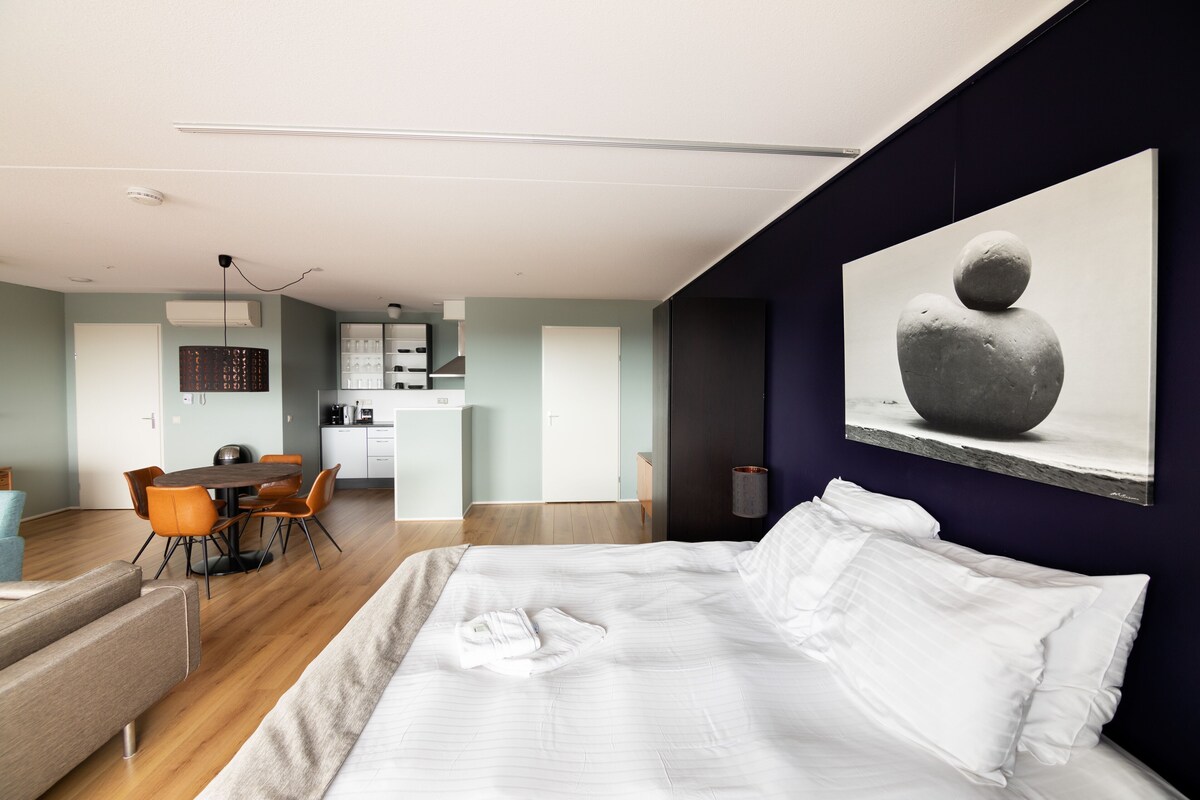 Una habitación de hotel moderna con una cama grande, una zona de comedor y una cocina al fondo.