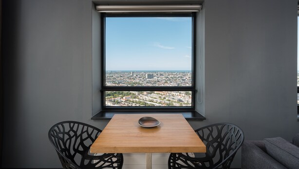 A room with a view of a cityscape through a large window, a wooden table with a bowl, and two black chairs. - The Penthouse (The Hague)
