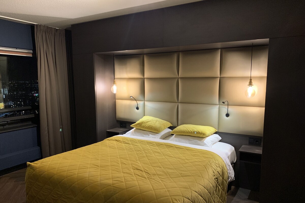 Habitación de hotel con una cama grande, cabecera con iluminación integrada, mesita de noche y vista nocturna de la ciudad.
