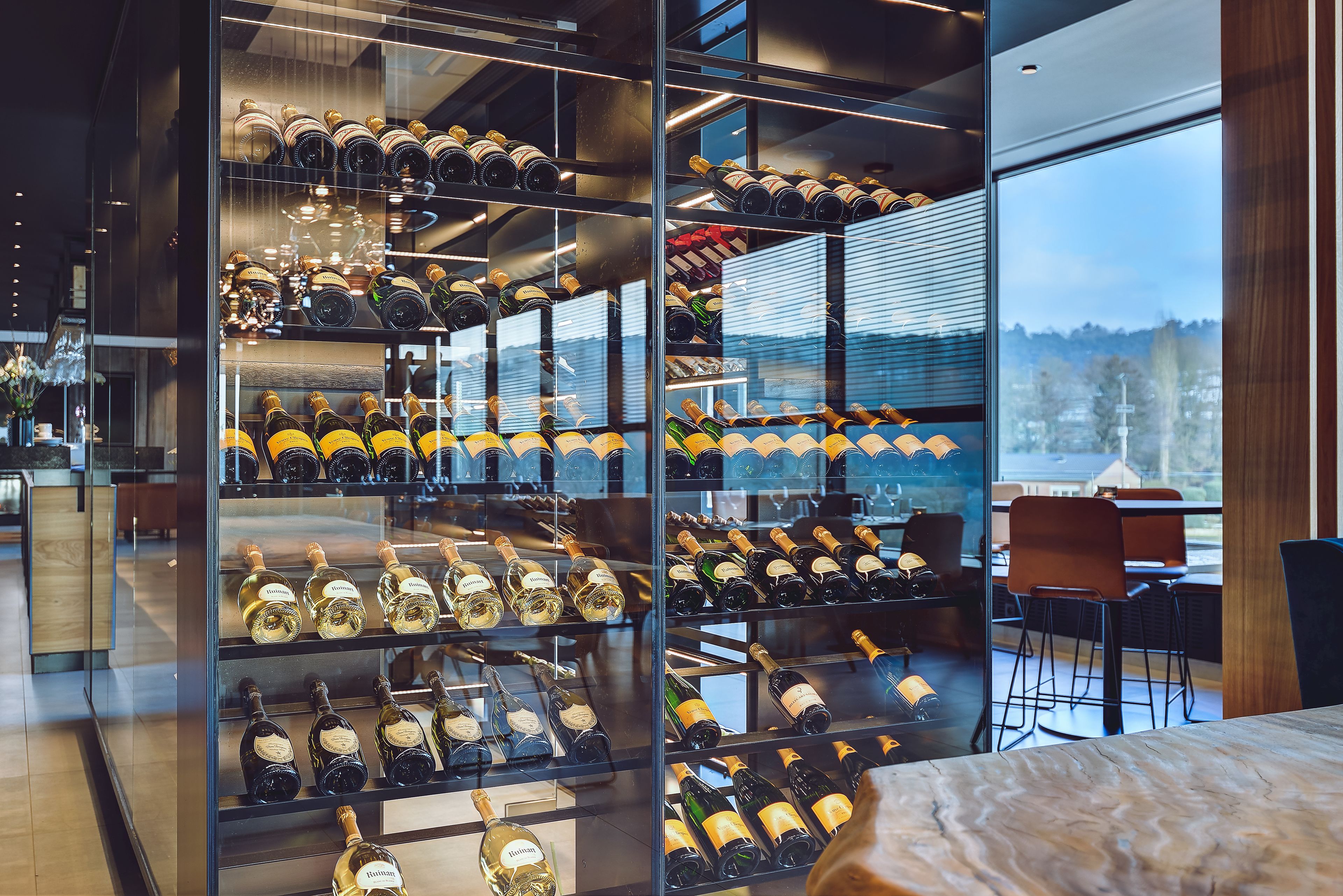 Una bodega con numerosas estanterías llenas de botellas de vino, un comedor con sillas y vistas a través de amplios ventanales.