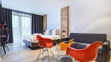 Ett modernt hotellrum med en säng, två röda fåtöljer, en grå soffa, ett litet bord med en flaska champagne och ett skrivbord med en lampa.