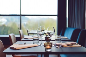 Una mesa de comedor con copas de vino, una vela y cubiertos, frente a una ventana con vista a árboles.