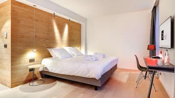 Ein modernes Hotelzimmer mit einer Wand in Holzoptik, einem Bett mit weißer Bettwäsche, einem Schreibtisch mit Lampe und einem Sessel.