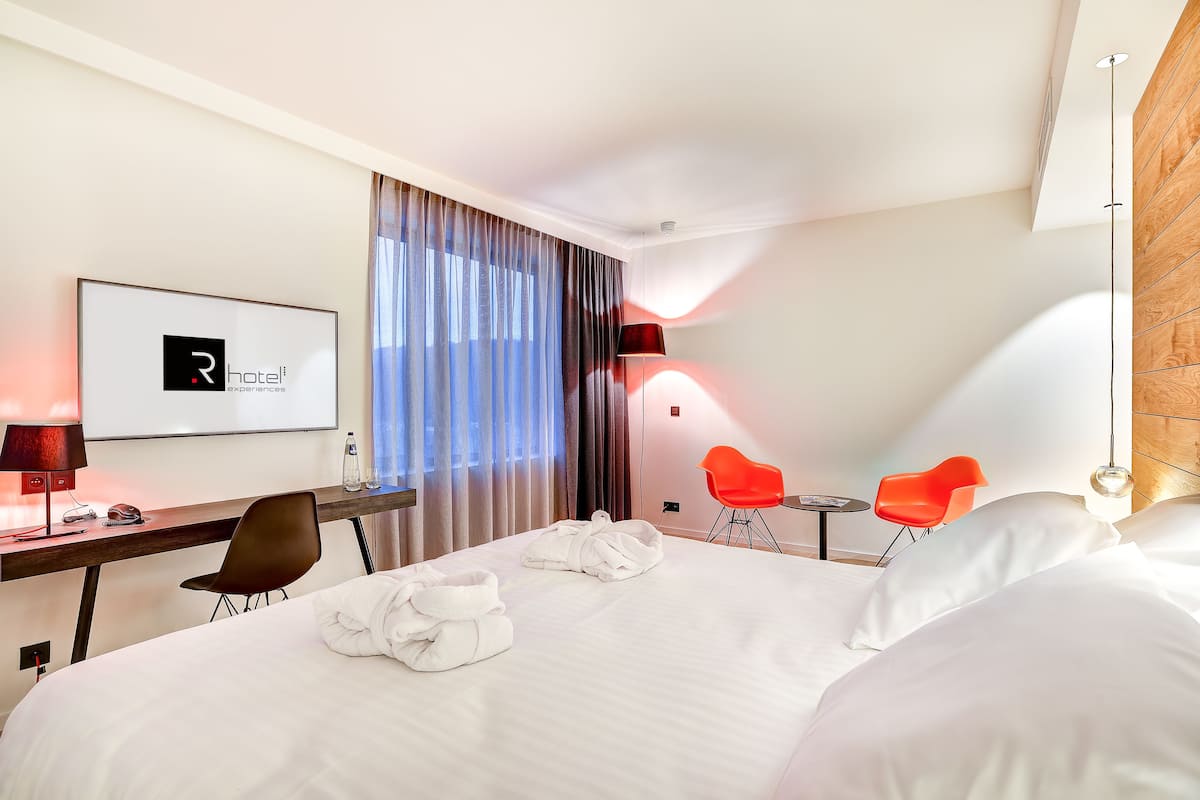 A hotel room with a bed, a desk with a computer, a chair, a red chair, and a TV displaying the hotel logo.
