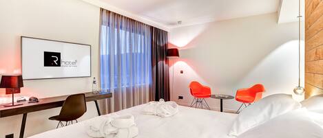 Habitación de hotel con una cama, un escritorio con computadora, una silla y una silla roja, además de un televisor mostrando el logotipo del hotel.
