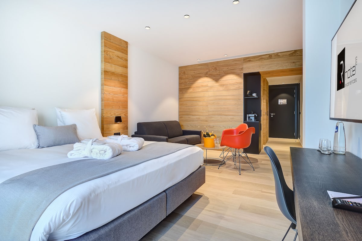 A modern hotel room with a large bed, a desk, a chair, and a small seating area.