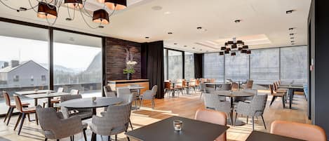Un comedor moderno con mesas redondas y sillas estampadas, ventanas amplias que ofrecen vistas al exterior y un techo con múltiples luminarias.