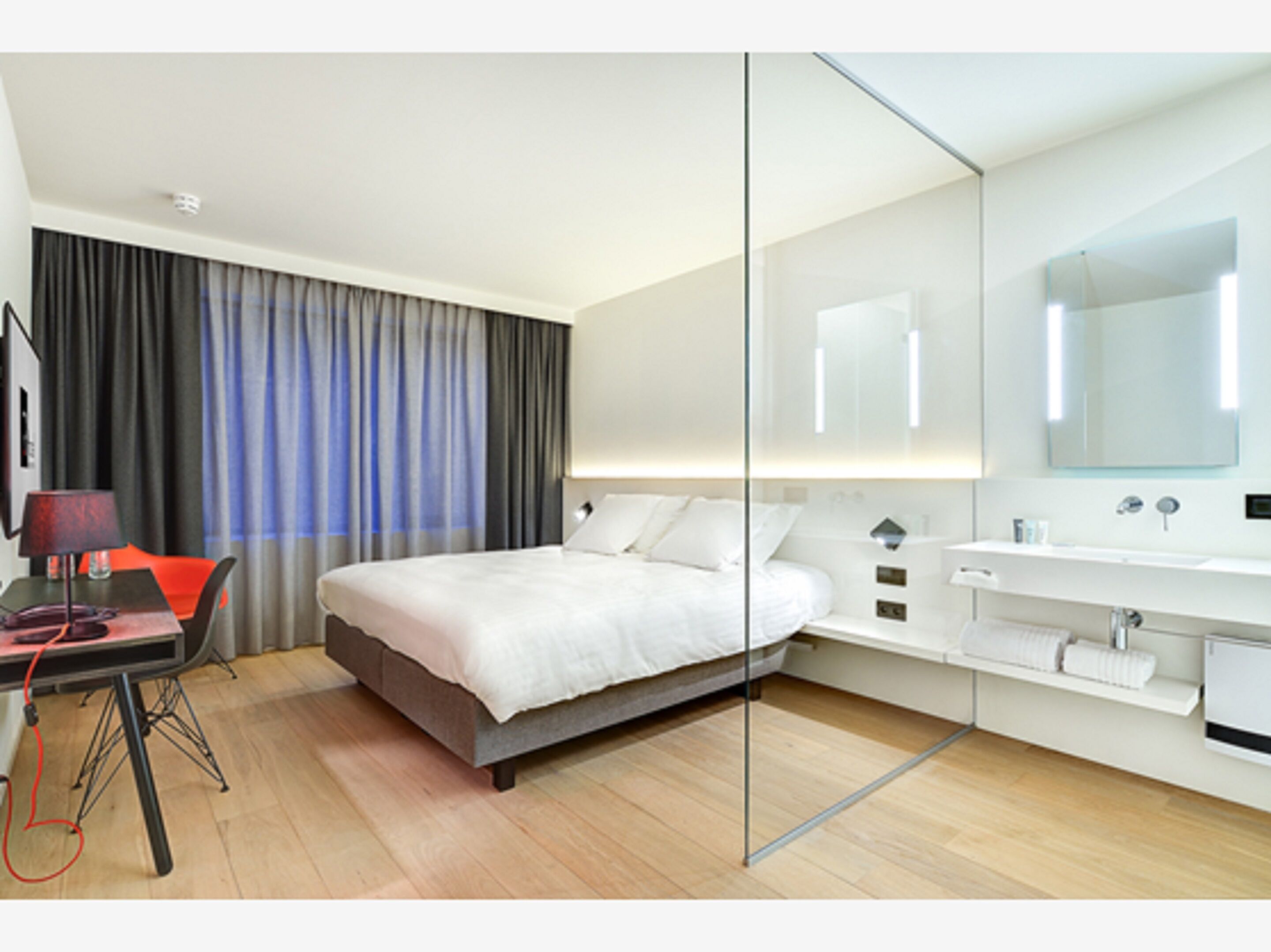 Una habitación de hotel moderna con una cama, un escritorio con lámpara, un espejo grande y un baño con lavamanos y espejo.