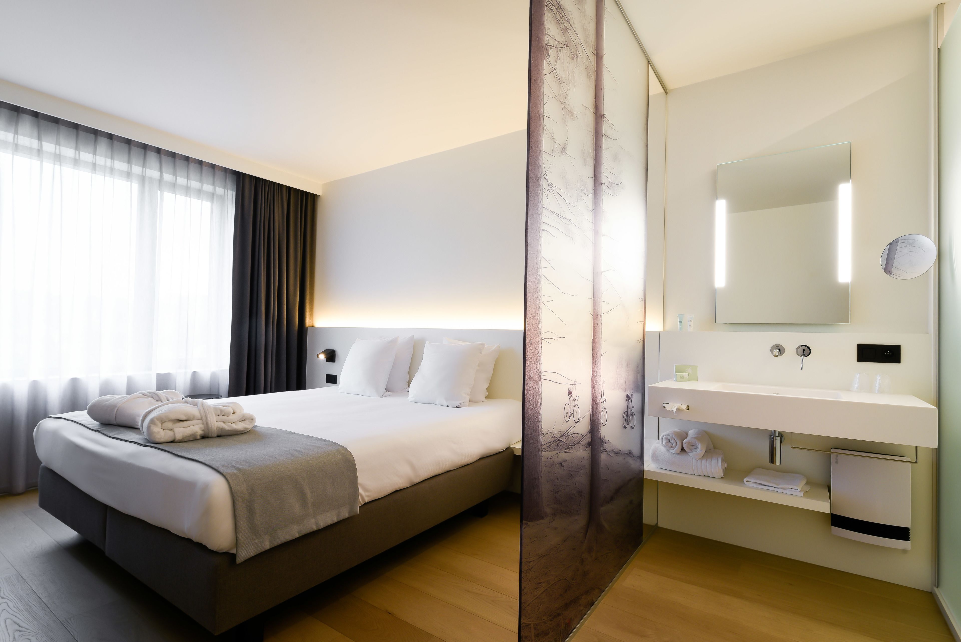 Une chambre d’hôtel moderne dotée d’un grand lit, d’une table de chevet, d’un miroir et d’une salle de bain avec un lavabo et un porte-serviettes.