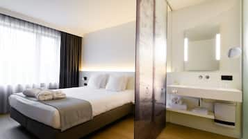 A modern hotel room with a large bed, a bedside table, a mirror, and a bathroom with a sink and towel rack.