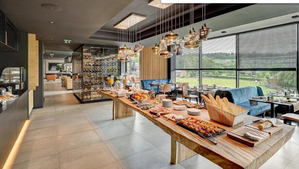 A modern restaurant with a long wooden counter, various food items, and a view of greenery through large windows. - R hotel experiences (Aywaille)