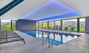 A modern indoor swimming pool with clear glass walls, lounge chairs, and a stone wall.
