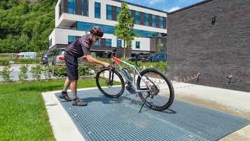 Uma pessoa ajustando uma bicicleta num tapete, em frente a um edifício com design moderno e um estacionamento ao fundo.