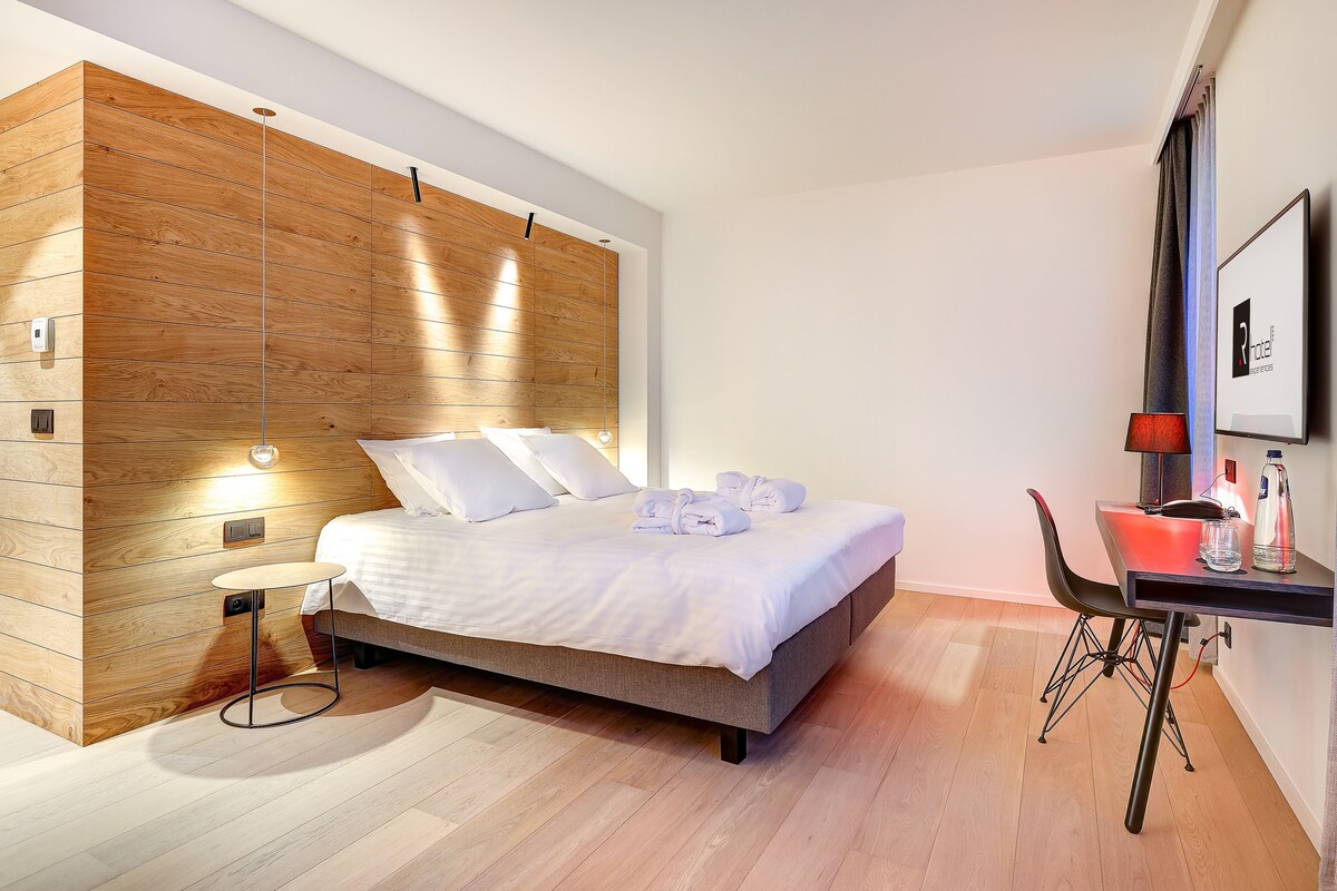 Una habitación de hotel moderna con cabecera de madera, cama bien hecha, una mesita redonda, escritorio con silla y un cuadro en la pared.